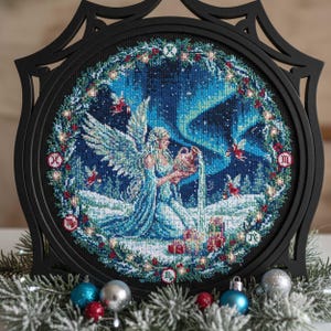 May include: A framed cross-stitch artwork featuring a celestial scene. The artwork depicts an angel pouring water, set against a backdrop of a starry night and aurora borealis. The frame is black and ornate, with decorative elements. The overall color palette includes blues, whites, and reds.