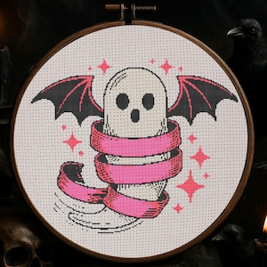 Puede incluir: Bordado de punto de cruz en un aro de madera con un fantasma blanco con alas de murciélago negras y una cinta rosa. Acentos de estrellas rosas rodean el fantasma. El fondo es oscuro.