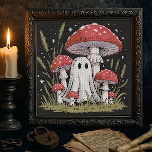 Op de afbeelding: Kruissteekkunst in een zwarte lijst met een grillige scène met rood-witte gestippelde paddenstoelen, een vriendelijk spook en hoog gras. Het kunstwerk heeft een donkere, gotische esthetiek, met een brandende kaars en een antiek slot op de voorgrond.