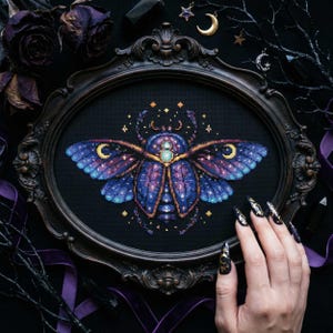 Könnte beinhalten: Ein Kreuzstich-Kunstwerk in einem ovalen Rahmen, das einen himmlischen Käfer mit blauen und violetten Flügeln darstellt, verziert mit Sternen und Monden. Der dunkle Rahmen ist verziert, vor einem schwarzen Hintergrund. Das Bild hat ein gotisches Thema.
