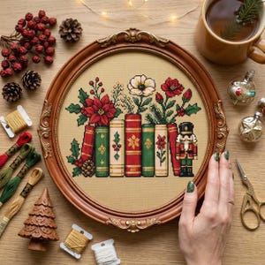 May include: A round wooden frame containing a cross-stitch embroidery of a row of books, a nutcracker, and festive flowers. The books are red, green, and cream with floral patterns. A white flower and red poinsettias sit atop the books.