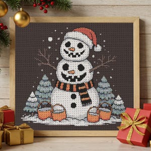 May include: Cross-stitch artwork of a snowman wearing a red hat and a black and orange scarf. The snowman has a smiling face and is surrounded by snow, trees, and buckets. The artwork is framed in a wooden frame.