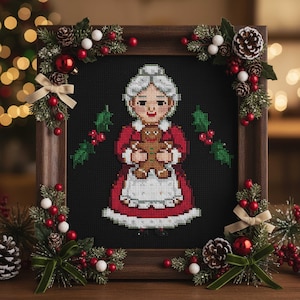 May include: Cross-stitch of a woman in a red and white dress holding a gingerbread man, framed in a wooden frame. The frame is decorated with greenery, red berries, white balls, and pine cones, creating a festive holiday theme.