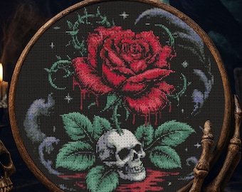 May include: Cross-stitch embroidery in a wooden hoop features a red rose with dripping blood, a skull, and green leaves against a black background. The design includes a crow and stars, creating a gothic theme.