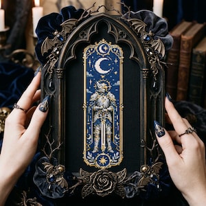 May include: A cross-stitch embroidery featuring a knight in silver armour holding a sword, set against a dark blue night sky with a crescent moon and stars. The piece is encased in an ornate, dark, arched frame decorated with bat wings and roses.