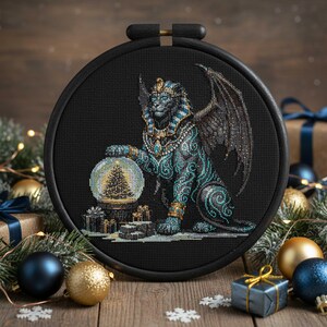 May include: A cross-stitch embroidery featuring a winged sphinx with teal and gold accents, holding a snow globe with a Christmas tree. The artwork is set in a black hoop, surrounded by Christmas ornaments and gifts on a wooden surface.