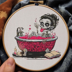 Puede incluir: Bordado de punto de cruz en un aro de madera que representa un esqueleto en una bañera cubierta de rosas y llena de líquido rosa. El esqueleto tiene la lengua fuera. Un libro y velas están al lado. El fondo es blanco.