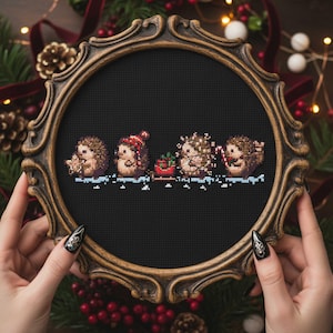 Può includere: Un disegno a punto croce con quattro ricci in una cornice decorativa. I ricci sono adornati con accessori festivi, tra cui un cappello di Babbo Natale e luci a stringa. Una piccola slitta rossa trasporta un regalo. La cornice è dorata e ornata.