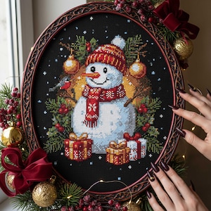 Könnte beinhalten: Ein gerahmtes Kreuzstichbild mit einem Schneemann mit roter Mütze und Schal, umgeben von Weihnachtsdekorationen und Geschenken. Der schwarze Hintergrund und der verzierte Rahmen erzeugen eine festliche Weihnachtsatmosphäre.