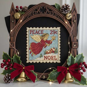 Puede incluir: Un bordado enmarcado con un diseño de sello postal vintage que presenta un ángel, las palabras "PEACE" y "NOEL", y el precio "29¢". El marco marrón está decorado con acebo, bayas, campanas y un lazo de terciopelo rojo. Luces de hadas envuelven el marco.