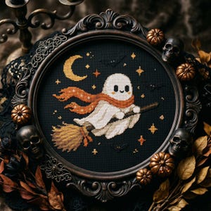 Puede incluir: Un bordado de punto de cruz circular que muestra un fantasma blanco con una bufanda naranja montado en una escoba sobre un fondo negro con una luna creciente amarilla, estrellas y murciélagos, en un marco decorativo de color marrón oscuro.