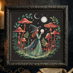 May include: Cross-stitch art in a dark, ornate frame. The artwork depicts a witch in a long green dress, holding a crystal ball, surrounded by red mushrooms and foliage. A crescent moon and full moon are in the night sky.