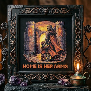 May include: A framed cross-stitch artwork with a gothic theme. The image depicts a figure in armor and a flowing cape, standing in a castle doorway. The text "HOME IS HER ARMS" is stitched in orange. The frame is black with ornate bronze accents.