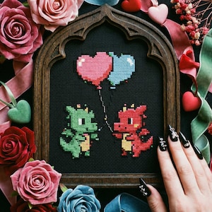 May include: Cross-stitch artwork showing two pixelated dragons with heart-shaped balloons. The dragons are green and red, on a black background in a wooden frame. Surrounded by roses, ribbons, and heart-shaped decorations.