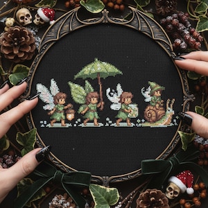 Op de afbeelding: Kruissteekkunst met vier feeën in groene outfits, met vleugels, die een lantaarn, een paraplu en een pot dragen. Eén fee rijdt op een slak. Het kunstwerk is ingelijst in een decoratieve, donkergekleurde lijst.