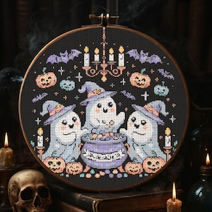 Puede incluir: Un bastidor de bordado de punto de cruz con un diseño de Halloween. Tres fantasmas amigables con sombreros de bruja rodean un caldero. Calabazas, murciélagos y velas decoran el fondo negro. El bastidor es de madera.