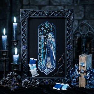 May include: A framed cross-stitch artwork featuring a figure in a blue hooded cloak holding a sword. The artwork is in a black frame with decorative embellishments. Spools of blue thread and lit candles are in the background, creating a gothic theme.