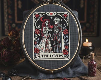 May include: A framed cross-stitch depicting a skeletal couple, surrounded by red roses, with the text "THE LOVERS." The artwork is set against a dark background, with a wooden frame. The overall theme is gothic and romantic.