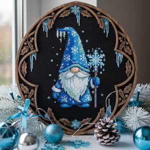 Puede incluir: Un bordado de punto de cruz de un gnomo de invierno en azul, sosteniendo un bastón de copo de nieve, sobre un fondo negro. La obra de arte está enmarcada en un marco ovalado de madera con bordes decorativos. También hay carámbanos y copos de nieve bordados.
