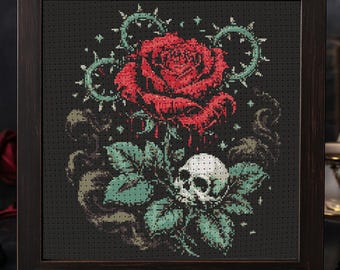 May include: Cross-stitch art in a dark wooden frame. The design features a large red rose, a skull, and green leaves, surrounded by a thorny vine on a black background. The artwork has a gothic theme.