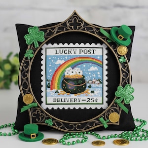 Puede incluir: Un diseño de punto de cruz con un arcoíris sobre una olla de oro, enmarcado con un marco decorativo de bronce. El texto "LUCKY POST" y "DELIVERY - 25¢" son visibles. El diseño está sobre un cojín negro, decorado con tréboles y monedas de oro.