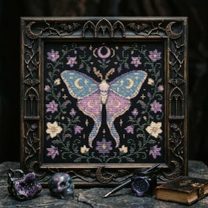 May include: A framed cross-stitch embroidery featuring a large moth with blue and purple wings, surrounded by floral vines and a crescent moon on a black background. The frame is dark, ornate, and decorated with small bats and moon motifs.