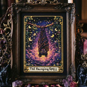 May include: Framed cross-stitch art featuring a bat hanging upside down, surrounded by moons and stars. The artwork is in a dark color palette with purple, yellow, and orange hues. The text "THE HANGING SPELL" is at the bottom. The frame is ornate and dark brown.