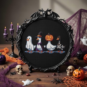 May include: A black framed cross-stitch featuring four ducks in Halloween costumes: a ghost, a witch, a jack-o'-lantern, and a skeleton. The frame has a decorative bat and skull design. The scene is set on a wooden table with Halloween decorations.