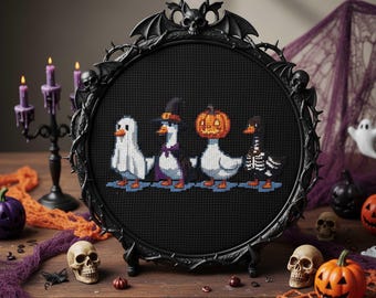 May include: A black framed cross-stitch featuring four ducks in Halloween costumes: a ghost, a witch, a jack-o'-lantern, and a skeleton. The frame has a decorative bat and skull design. The scene is set on a wooden table with Halloween decorations.
