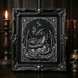 May include: Black framed cross-stitch art depicting a dragon and skulls. The gothic frame has ornate corners and a skull and crossbones detail. A candle and skull ornament are visible in the background.