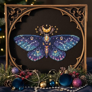 May include: Cross-stitch art of a moth with blue and purple wings, a yellow crescent moon, and star details. The artwork is framed in a dark wooden frame with ornate detailing. Decorative elements include pine needles, ornaments, and string lights.