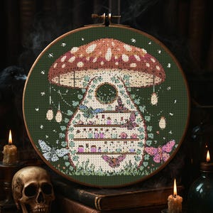 May include: Cross-stitch embroidery in a wooden hoop. The design features a mushroom with a dotted cap, a shelf filled with jars, and butterflies. The background is dark green, with hanging lights and a wooden frame.