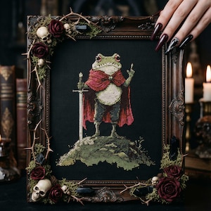 以下が含まれることがあります： 黒い背景に、赤いマントを着て剣を持ったカエルのクロスステッチアート。暗い木製のフレームは、頭蓋骨、バラ、とげで飾られています。カエルは緑の丘の上に立っています。