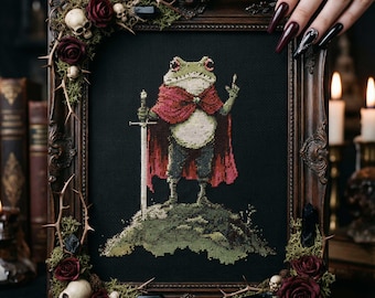 May include: Cross-stitch artwork in a dark wooden frame, depicting a frog in a red cape with a sword. The frame is decorated with skulls, roses, and thorns. The frog stands on a green hill against a black backdrop.