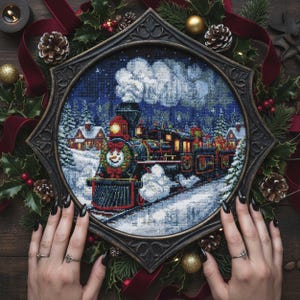 May include: A framed cross-stitch depicting a festive winter scene with a steam train. The train is decorated with holiday lights and a snowman face. The frame is surrounded by Christmas decorations, including holly, pine cones, and red ribbon.