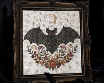 Puede incluir: Arte de punto de cruz enmarcado con un murciélago negro con ojos rojos, una luna creciente y estrellas. El murciélago está sobre una corona floral con colores otoñales. El marco es ornamentado y de color bronce oscuro.