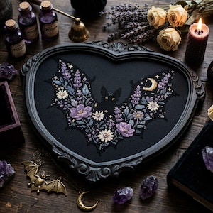 以下が含まれることがあります： 紫と白の花柄の翼を持つコウモリと三日月が刺繍された、ハート型の黒いフレーム。コウモリは暗い毛並みと輝く目をしており、ラベンダーの小枝と小さな花々に囲まれているクロスステッチの刺繍作品。