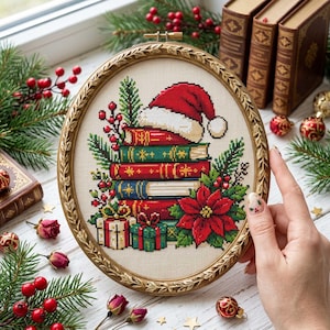 May include: An oval embroidery hoop containing a cross-stitch design of a stack of books topped with a Santa hat, surrounded by pine branches, red berries, a poinsettia, and two wrapped gifts, all set against a light fabric background.