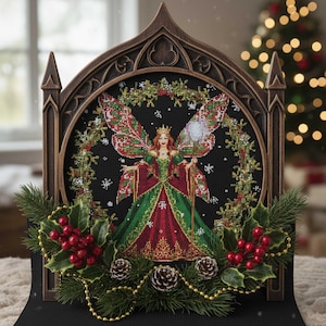 Może przedstawiać: Wyszywana krzyżykami bożonarodzeniowa wróżka w dekoracyjnej ramie. Wróżka ma na sobie czerwoną i zieloną suknię i pasujące skrzydła. Rama jest brązowa z gotyckim łukiem. Ostrokrzew, szyszki i jagody zdobią podstawę.