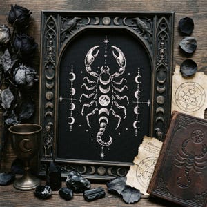 May include: A framed black and white cross-stitch depicting a scorpion, with crescent moons and stars. The dark wood frame has gothic detailing. The image includes a chalice, crystals, and a leather-bound book with alchemical symbols.