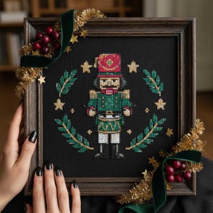 May include: A framed cross-stitch of a nutcracker soldier in green and red, set against a black backdrop. The design is encircled by a wreath of green leaves and gold stars. The dark wood frame is adorned with gold tinsel and a green velvet ribbon.