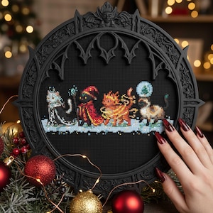 May include: A black, gothic-style frame with a cross-stitch design featuring four cartoon cats in festive attire. The cats are set against a snowy landscape, with a Christmas tree in a snow globe. Decorative Christmas ornaments are in the foreground.