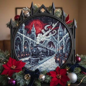 Könnte beinhalten: Ein Kreuzstich-Kunstwerk, das eine Winterszene mit einem Zug, Gebäuden und einem roten Mond darstellt. Das Kunstwerk ist in einem schwarzen Rahmen im gotischen Stil gerahmt und mit Weihnachtsschmuck, roten Weihnachtssternen und Tannenzweigen verziert.