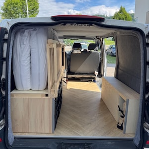 Op de afbeelding: Interieur van een witte camperbus. De bus is voorzien van op maat gemaakte houten meubels, waaronder een bedplatform, opbergruimtes en een bank. De vloer heeft een licht houtnerfpatroon. Twee witte matrassen zijn zichtbaar.