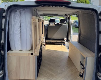 May include: Interior view of a white van converted for camping. The van features custom wooden cabinetry, including a bed platform, storage units, and a bench. The flooring has a light wood grain pattern. Two white mattresses are visible.