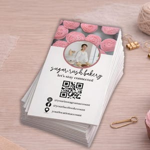 May include: A stack of white business cards with the text "sugar rush bakery" and "let's stay connected". The cards feature a QR code, social media handles, and a photo of baked goods. Pink cupcakes and gold paper clips are also visible.