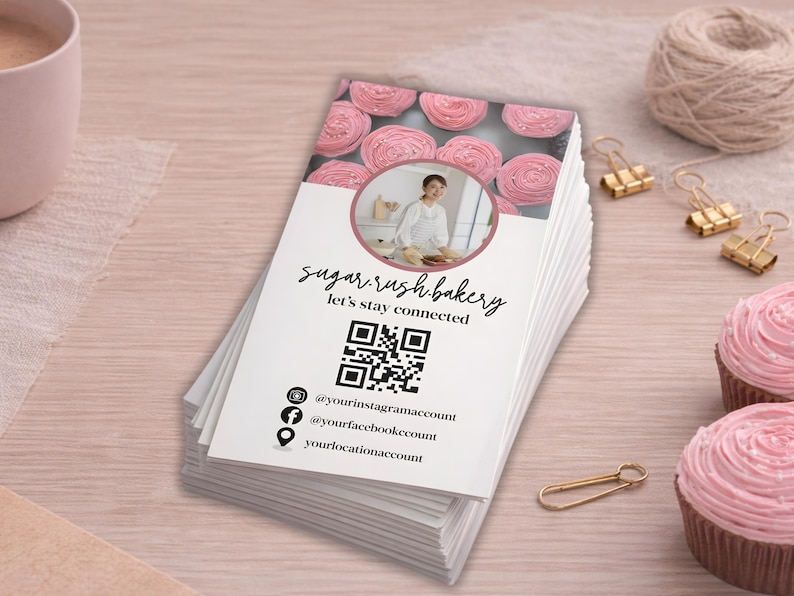 May include: A stack of white business cards with the text "sugar rush bakery" and "let's stay connected". The cards feature a QR code, social media handles, and a photo of baked goods. Pink cupcakes and gold paper clips are also visible.