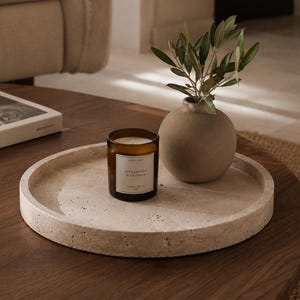 Pode incluir: Uma bandeja redonda de pedra bege porosa contendo uma vela de vidro castanho com um rótulo branco com a inscrição Bergamotto di Calabria e um pequeno vaso de cerâmica bege mate com um ramo verde de folhas pontiagudas.