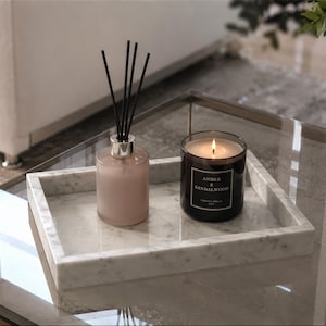 May include: A rectangular white marble tray holding a light pink reed diffuser with black reeds and a dark brown glass candle jar with a black label reading Amber & Sandalwood. The candle has a lit wick and sits next to the diffuser on the tray.