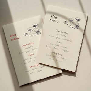 May include: Two off-white menu cards with the text "the menu" in red and black. The cards feature a simple illustration of wine glasses and food. The menu lists appetizers, mains, and desserts.